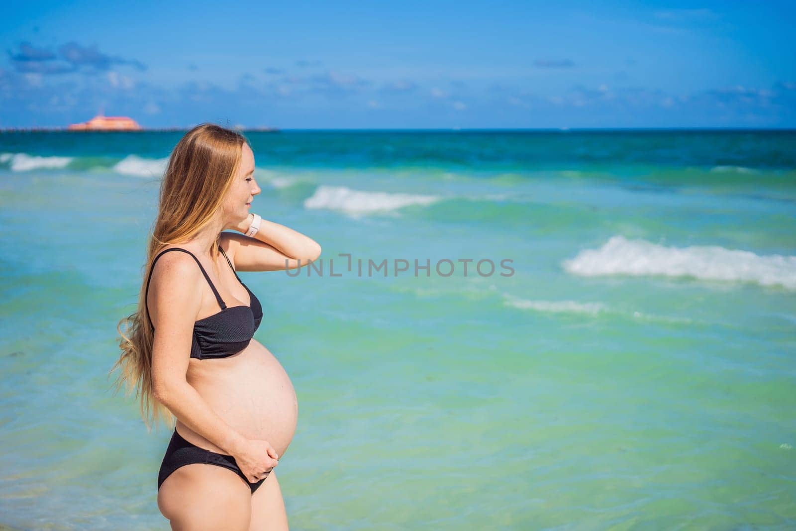 Radiant pregnant woman in a swimsuit, amid the stunning backdrop of a turquoise sea. Serene beauty of maternity by the shore.
