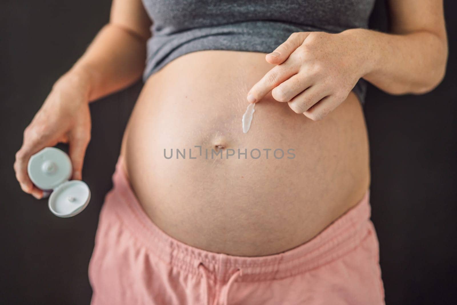 Positive young pregnant woman in comfortable homewear applying belly butter on her big tummy, woman belly by galitskaya