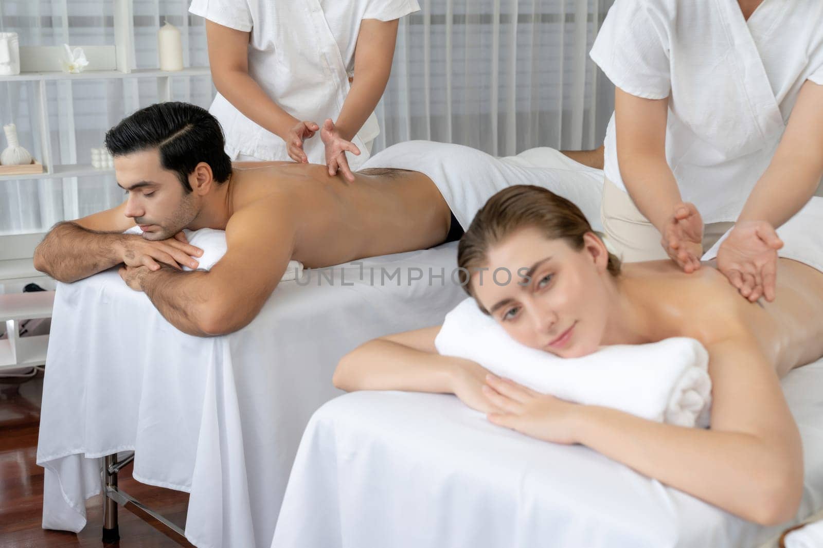 Caucasian couple customer enjoying relaxing anti-stress spa massage and pampering with beauty skin recreation leisure in day light ambient salon spa at luxury resort or hotel. Quiescent