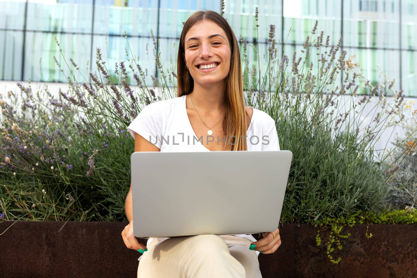 Front view of young female university student sitting on bench using laptop looking at camera. Female university student doing homework in campus outdoors. Education and technology concepts.