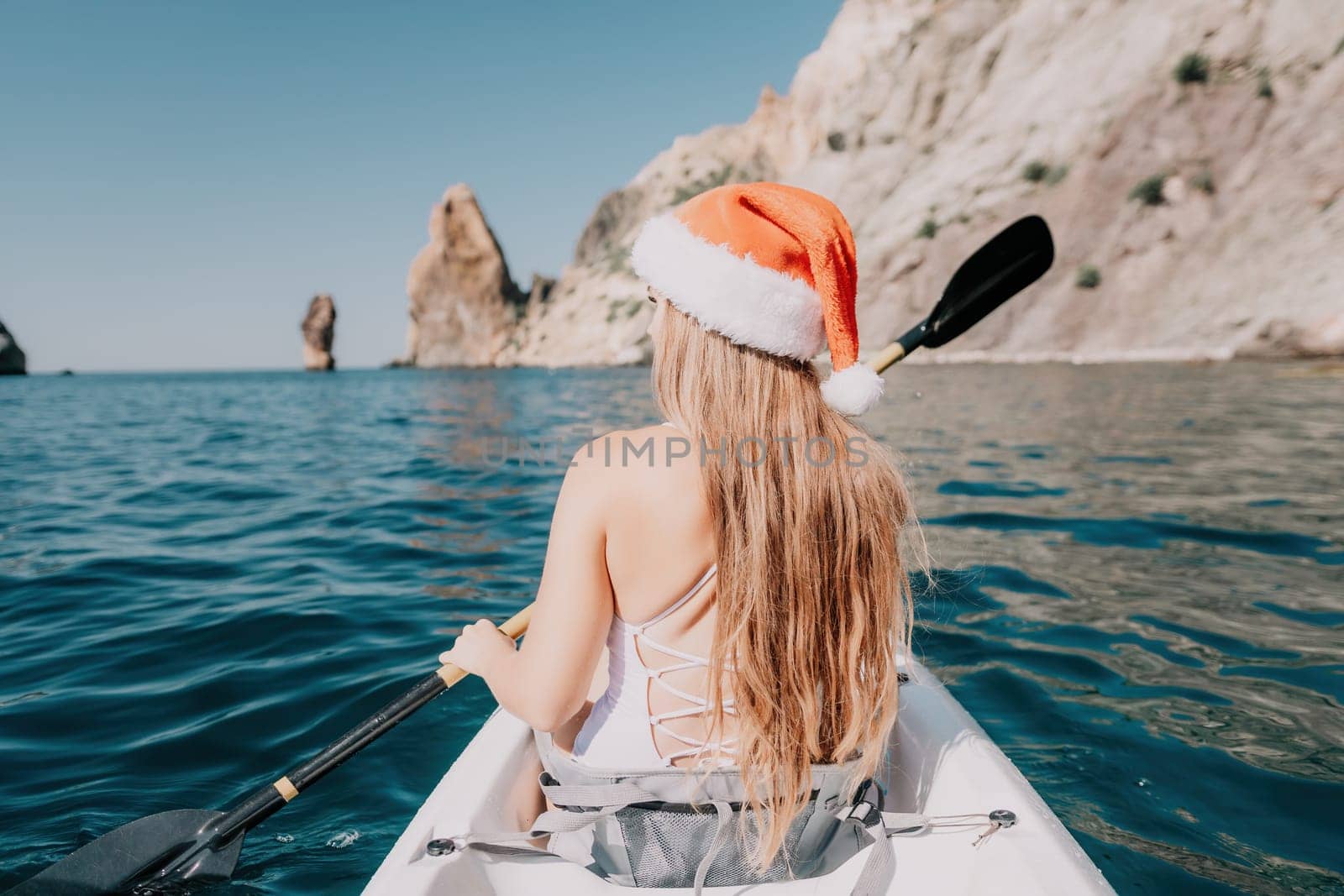 Woman in kayak back view. Happy young woman in Santa hat floating in kayak on calm sea. Summer holiday vacation and cheerful female people relaxing having fun on the boat. by panophotograph