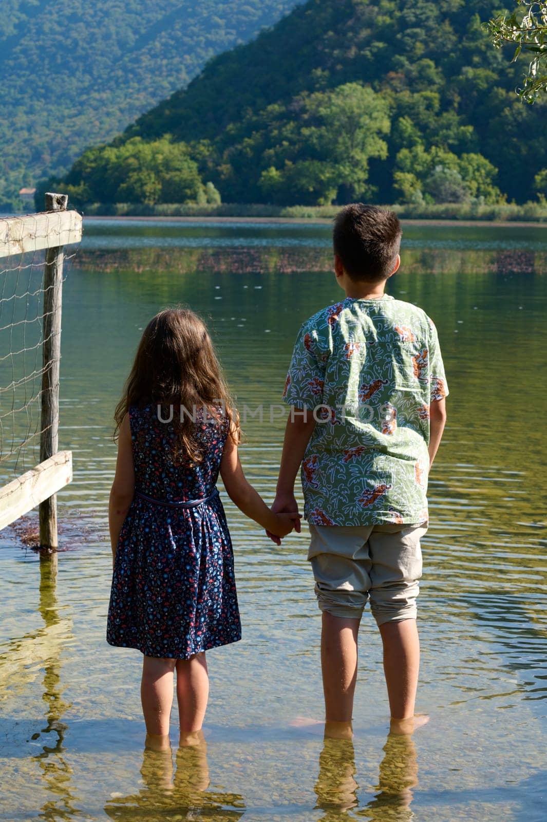 Two adorable kids, boy and girl holding hands, admiring the beautiful landscape of Segrino lake, Italian Alps background by artgf