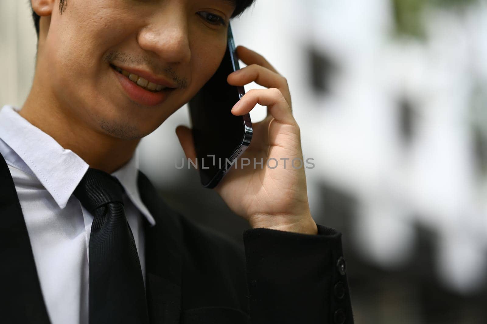 Millennial businessman in black suit having pleasant phone conversation while standing outside office building by prathanchorruangsak