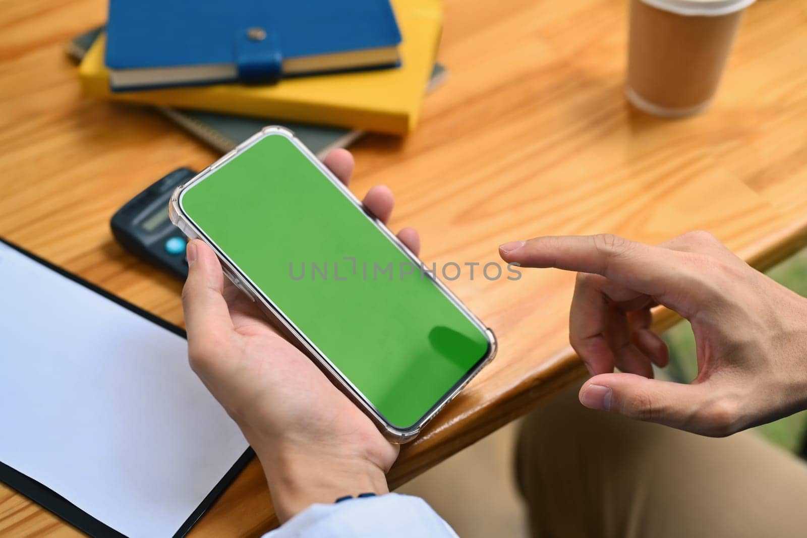 An unrecognizable man using smartphone with green screen on wooden office desk by prathanchorruangsak