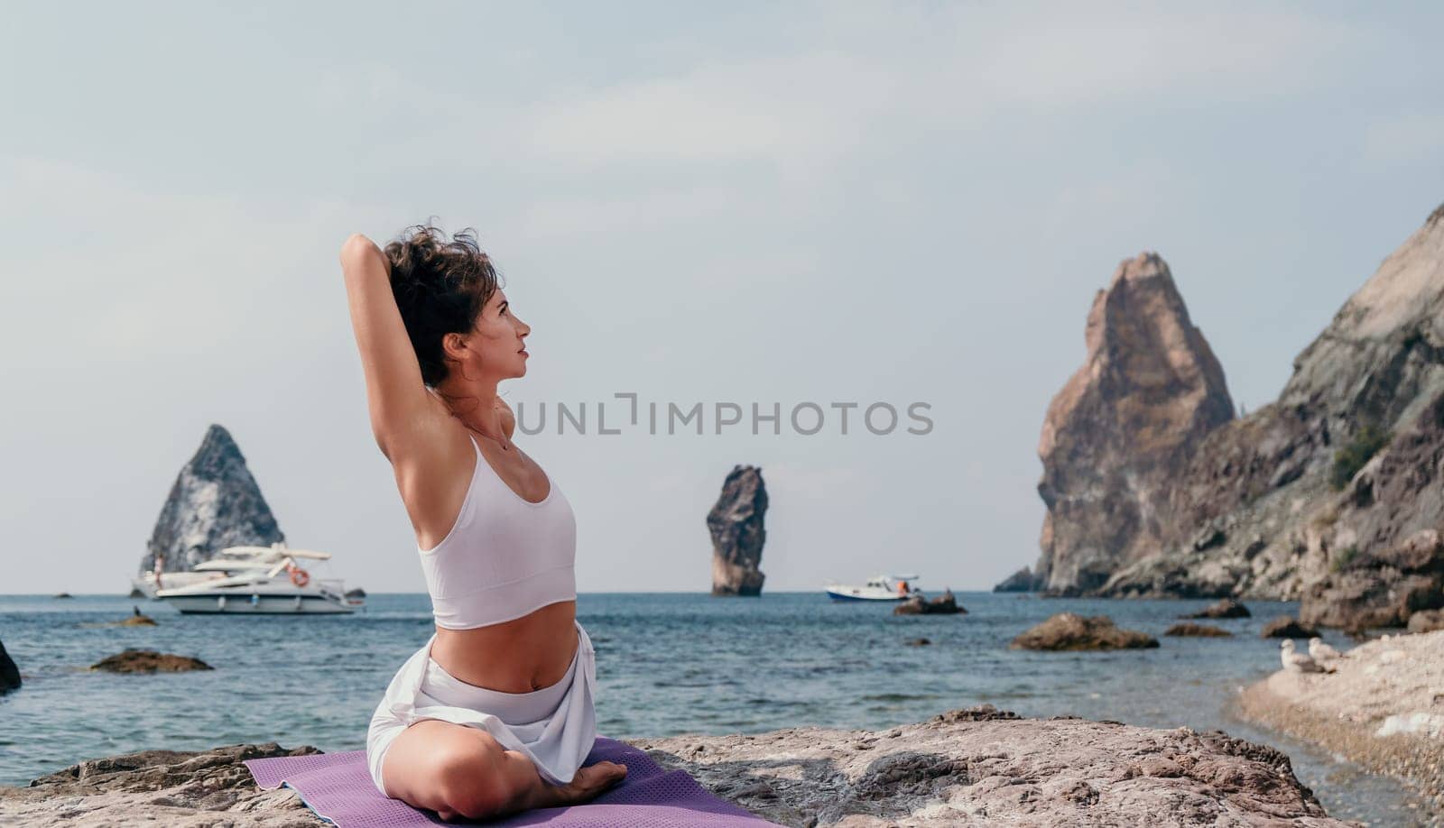 Fitness woman sea. A happy middle aged woman in white sportswear exercises morning outdoors on a beach with volcanic rocks by the sea. Female fitness pilates yoga routine concept. Healthy lifestyle. by panophotograph