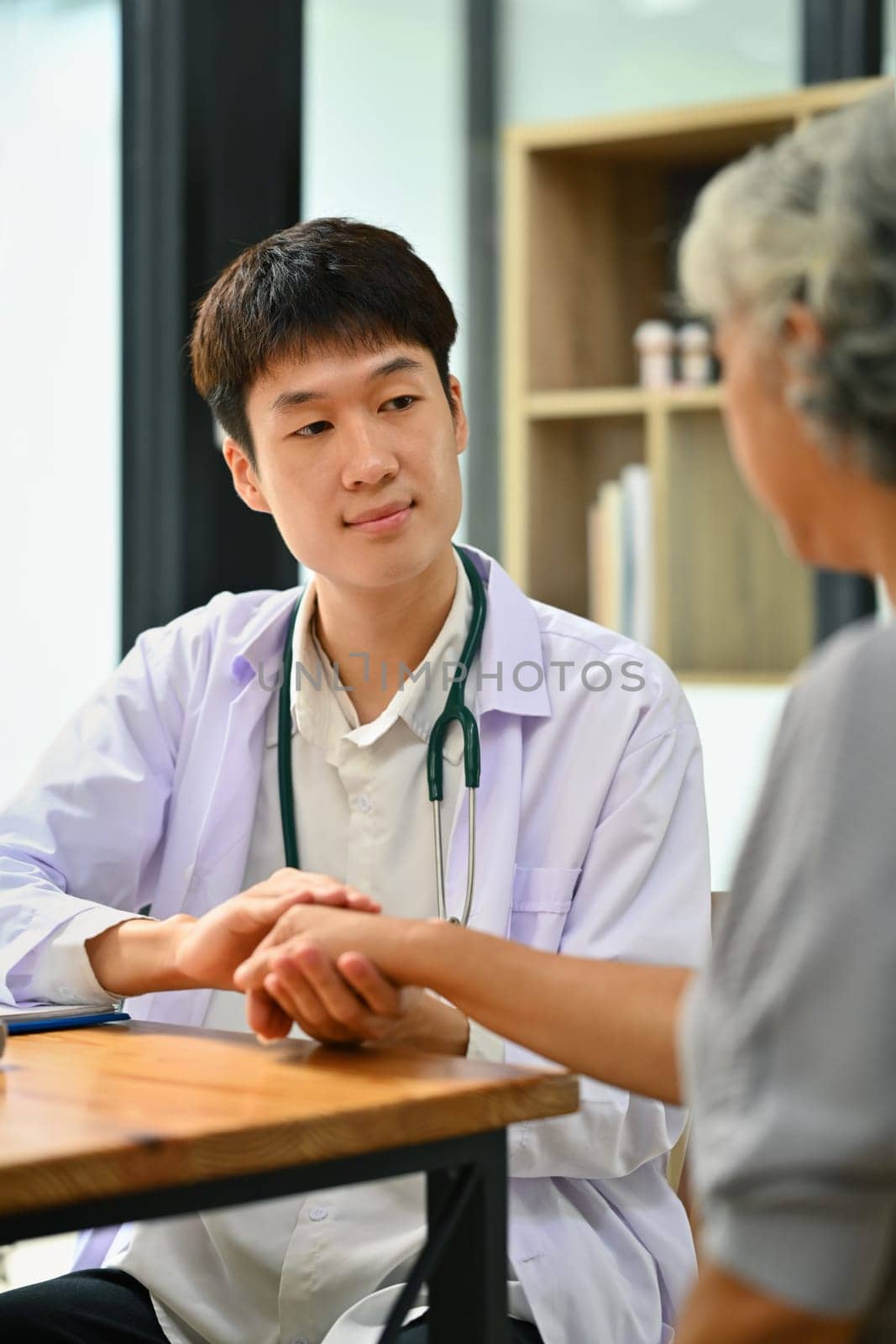 Caring asian male physician holding mature patient hands comforting and supporting at medical appointment.