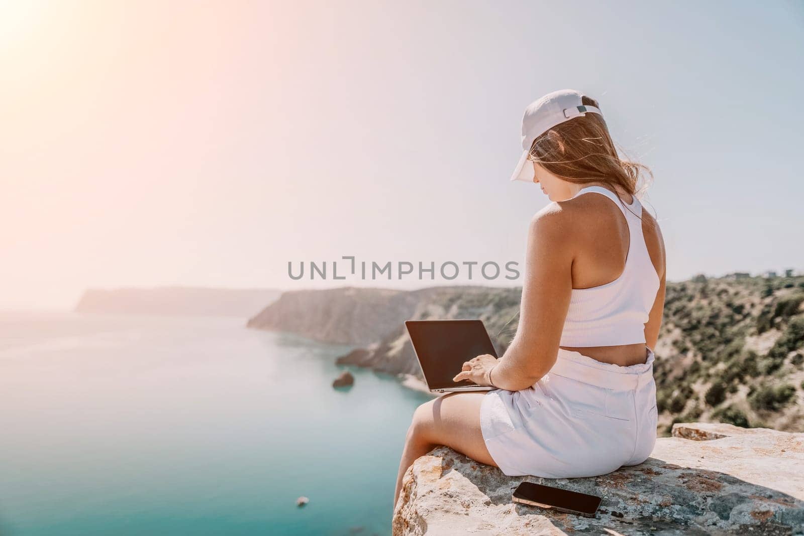 Digital nomad, Business woman working on laptop by the sea. Pretty lady typing on computer by the sea at sunset, makes a business transaction online from a distance. Freelance, remote work on vacation by panophotograph