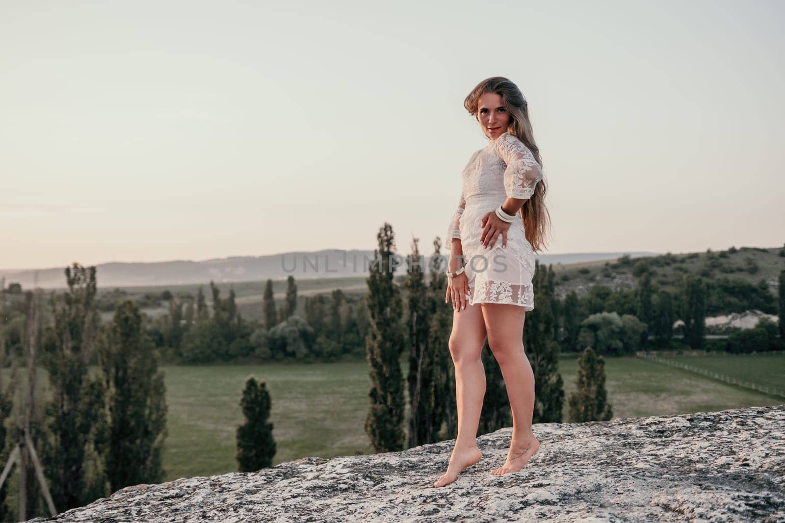 Happy woman in white boho dress on sunset in mountains. Romantic woman with long hair standing with her back on the sunset in nature in summer with open hands. Silhouette. Nature. Sunset. by panophotograph