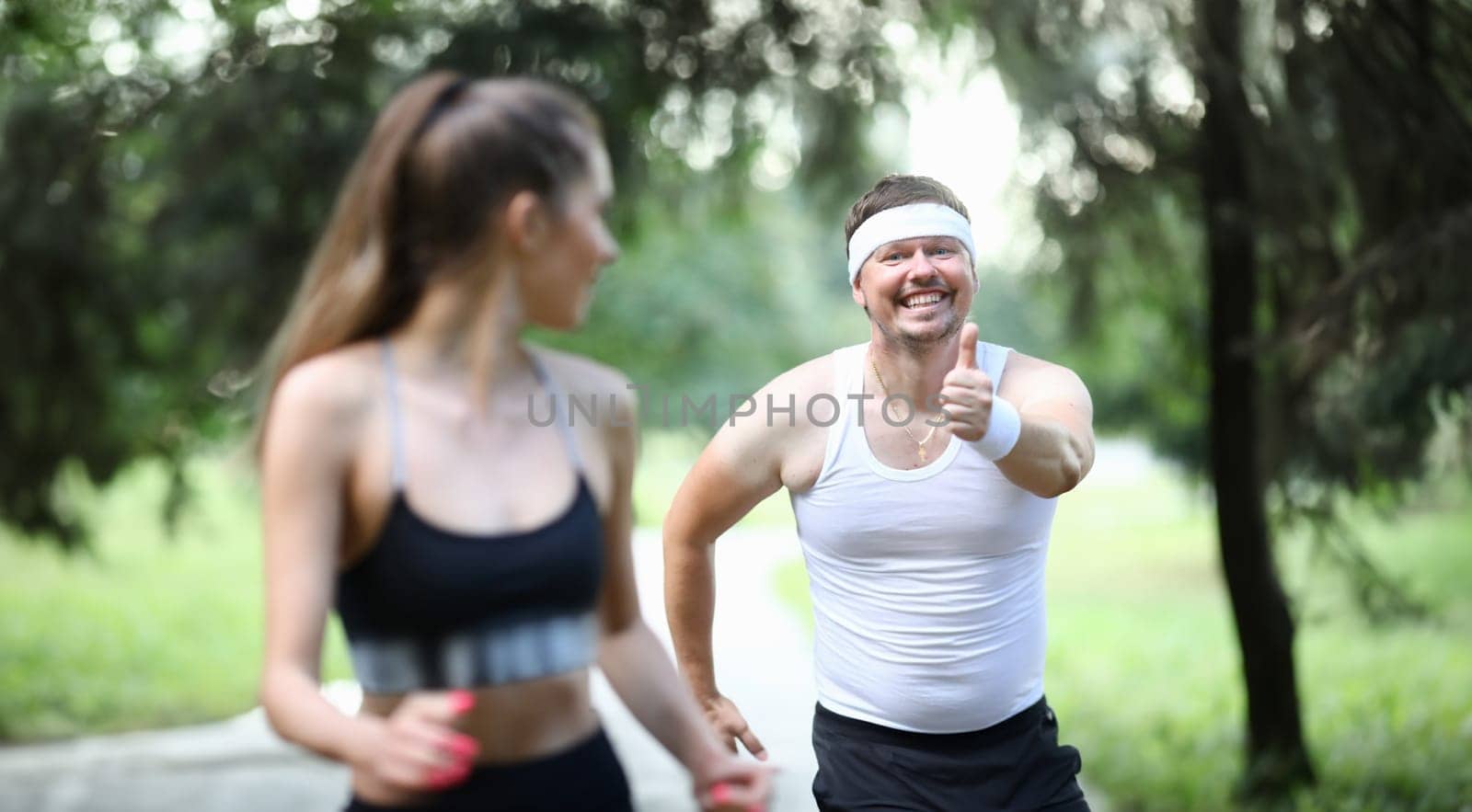 Portrait of couple training together in park. Man making funny faces. Middle-aged male showing thumbs up and running in sportswear. Sport and active lifestyle concept