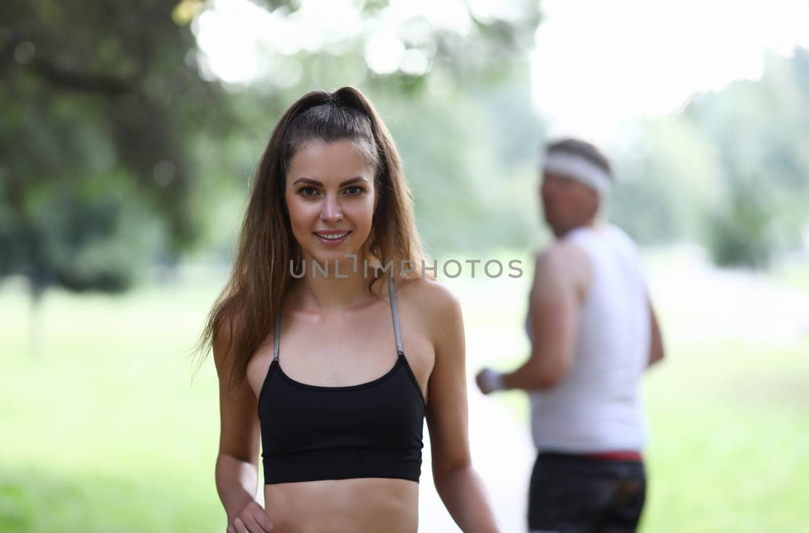 Portrait of beautiful fit female posing on camera outdoors. Morning park run for strong body. Man watching pretty girl from back. Sport and active lifestyle concept