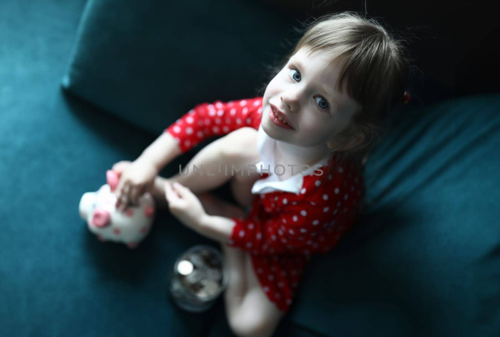 Top view carefree kid sitting on cozy sofa at home. Smiling beautiful girl wearing funny slippers. Child in pretty red dress in small dots. Childhood and happiness concept