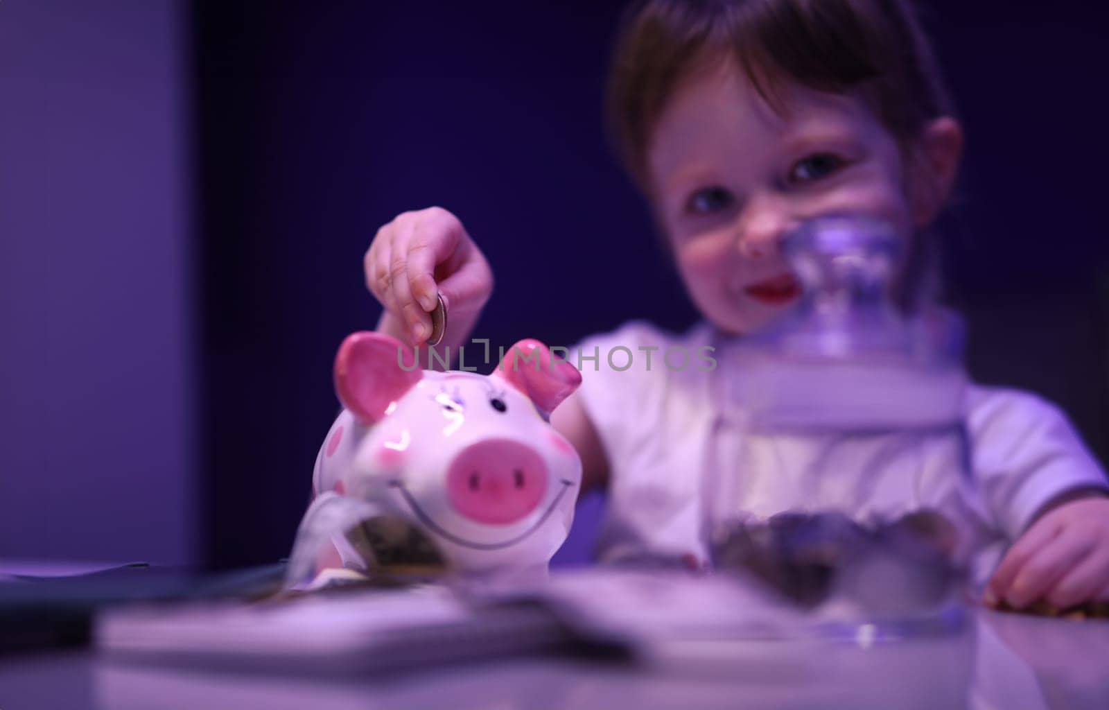 Portrait of lovely child putting coin in pink piggy bank. Smiling charming girl saving up money for future. Pretty kid holding cash. Childhood and happiness concept