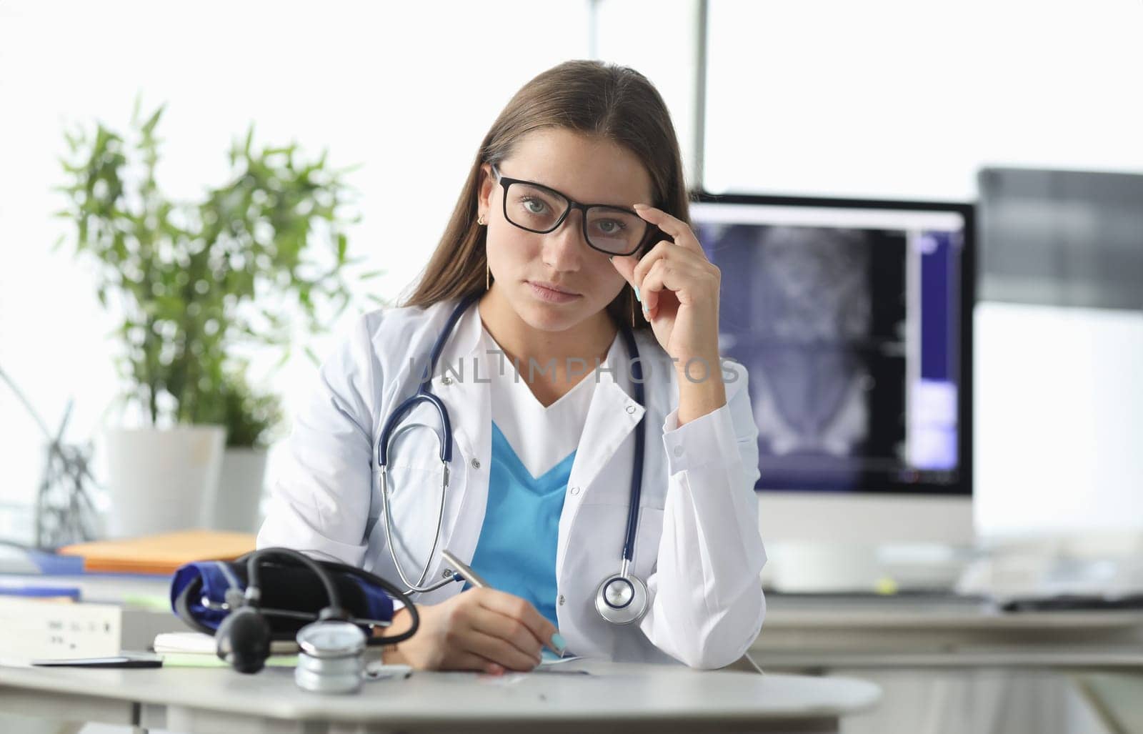 Portrait of young practitioner in glasses at workplace. Tonometer lying on table. Doctor office interior on background. Healthcare and medicine concept