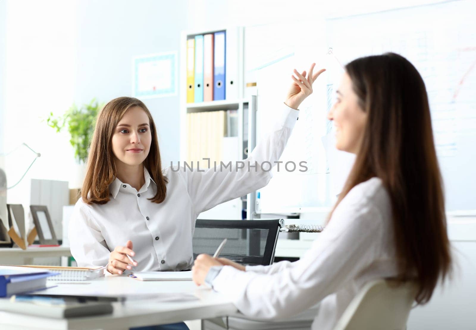 Portrait of businesswoman pointing with tender hand. Joyful female sitting at workplace with colleague and discussing important charts in significant paper tablet. Accounting office concept
