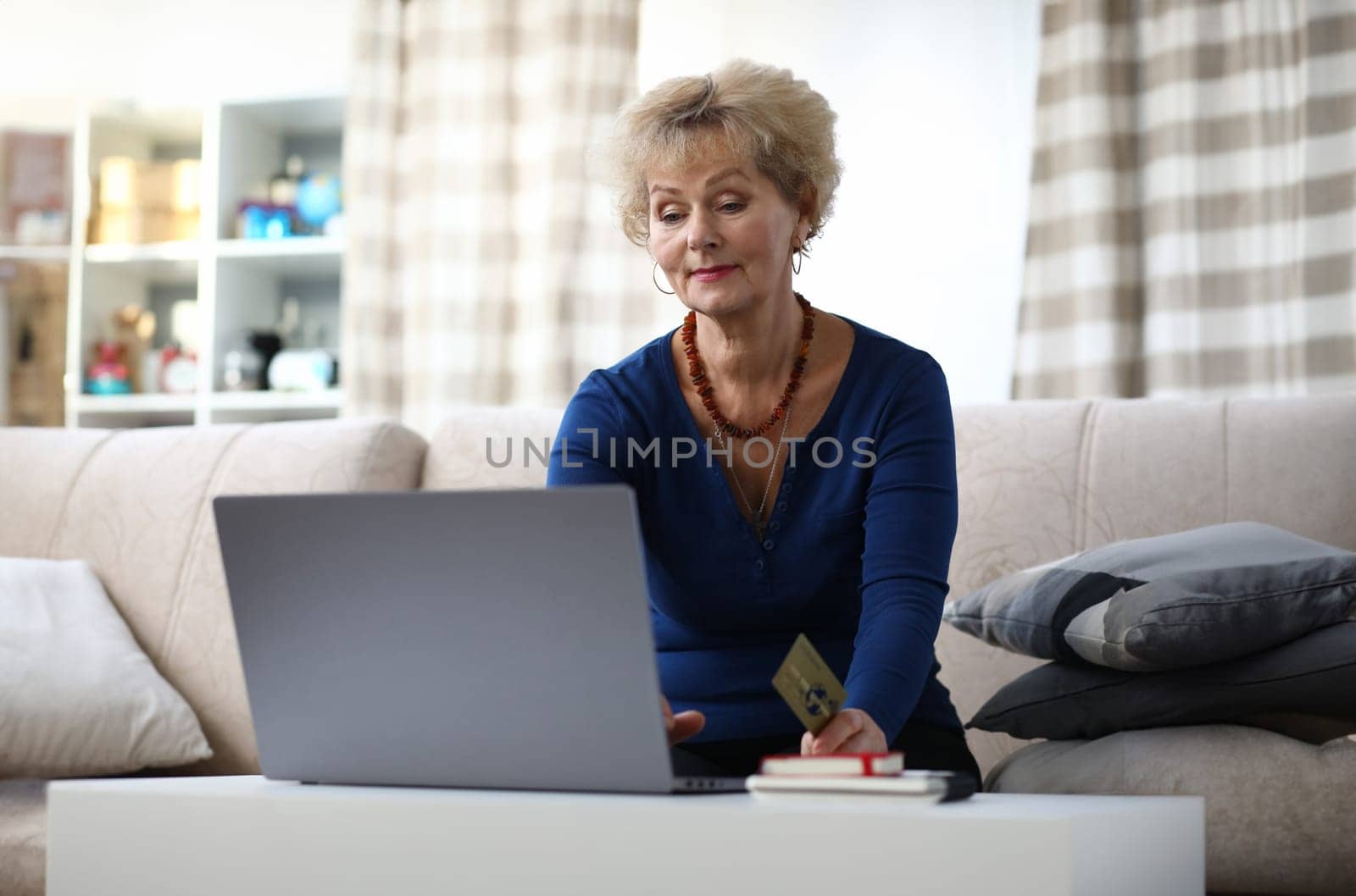 Portrait of beautiful elderly woman sitting on sofa at home. Charming female shopping via internet using laptop. Lady holding credit card. Modern technology and spare time concept