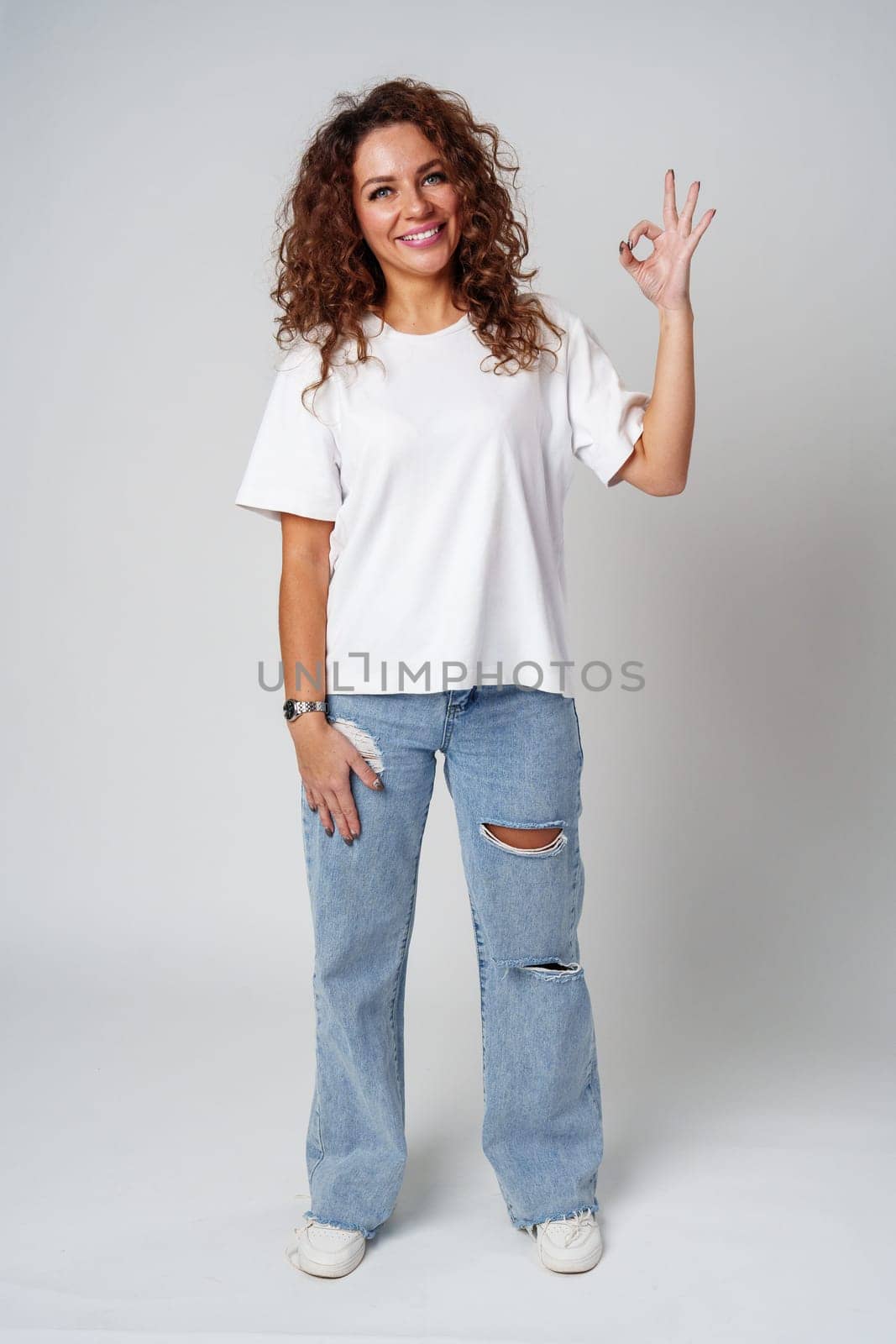 Cheerful young woman in a white shirt and ripped jeans making an okay gesture in studio