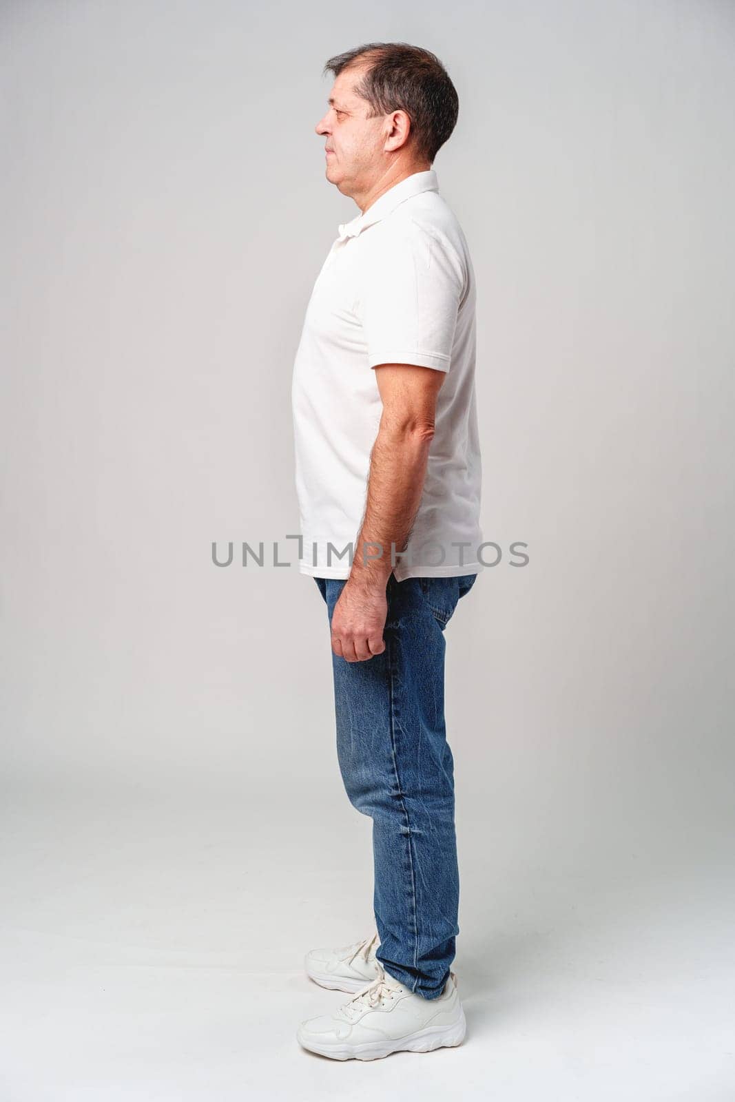 Mature man standing sideways in casual attire against a plain background