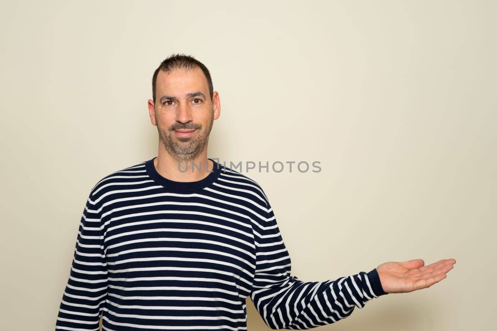 A bearded Hispanic man in his 40s wearing a striped sweater extending his hand to the side in a gesture of offering or outstretching, holding it palm forward. Isolated on beige background
