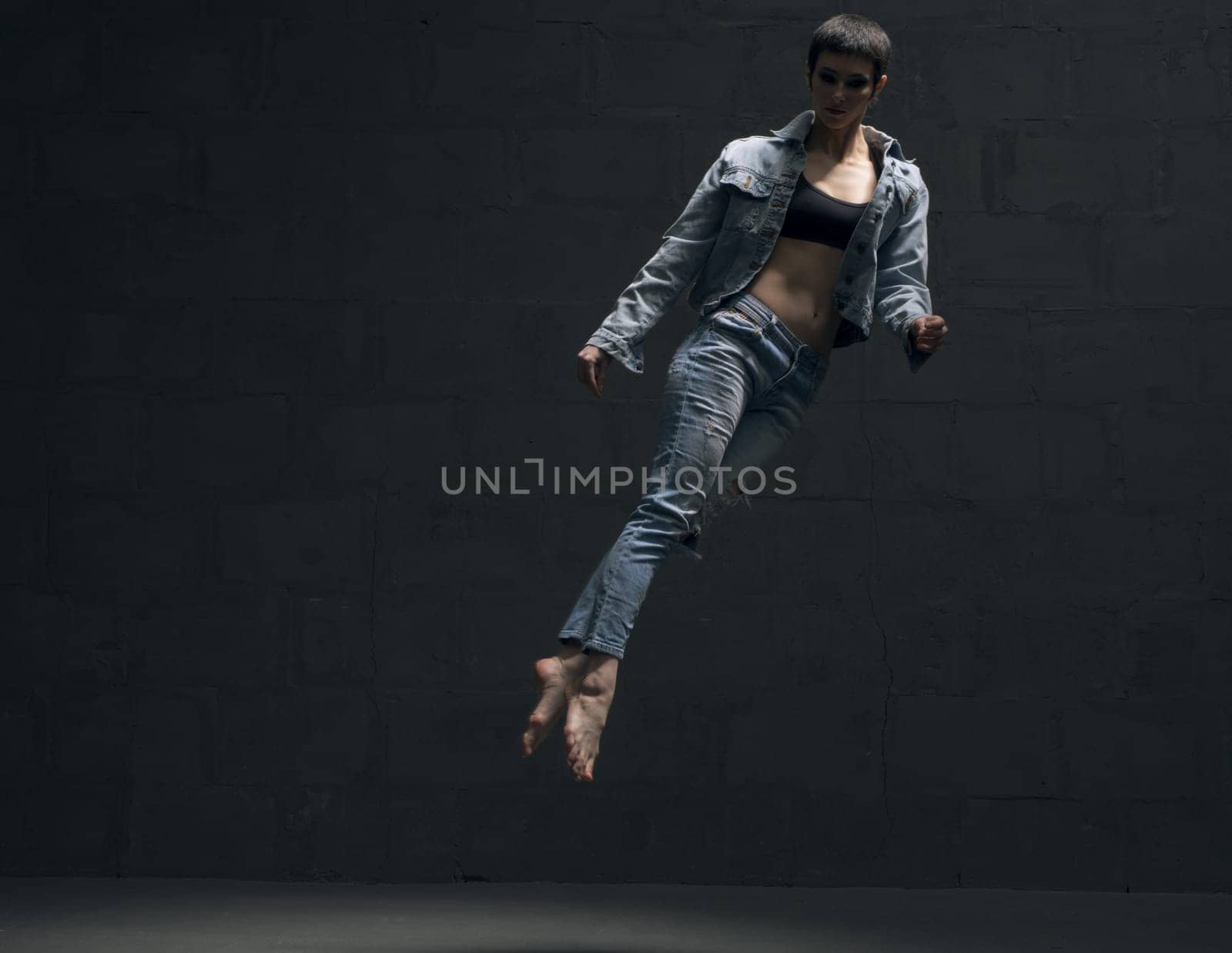 Girl with shot haircut in jeanswear jumping gracefully in dark room full-length shot