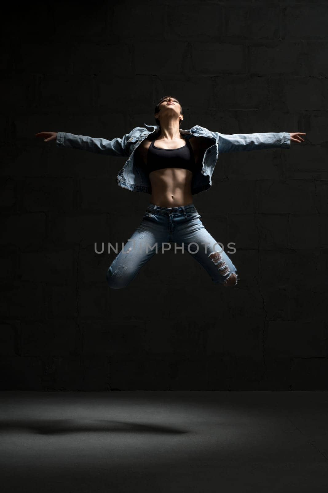 Ballet dancer with shot haircut in unbuttoned jeans shirt jumping gracefully in dark room