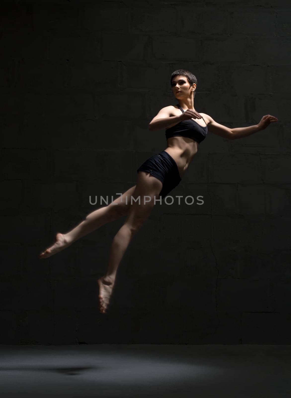 Ballet dancer with shot haircut in top and shorts jumping gracefully in dark room