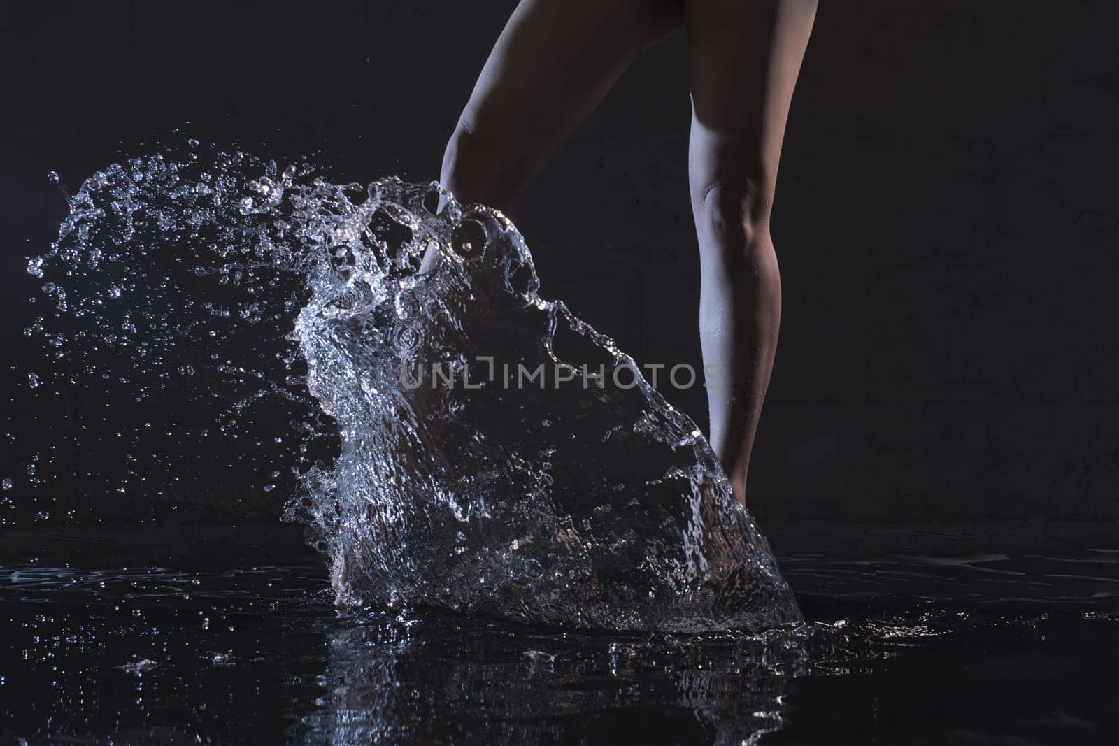 Barefoot slender woman dancing in water in dark room cropped shot