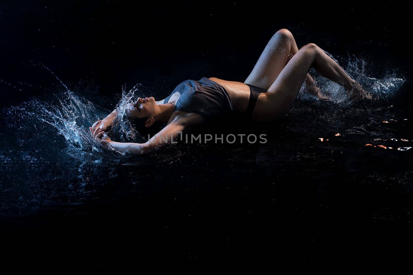 Sexy girl wearing top and strings lying in water in the dark room surrounded by splashes