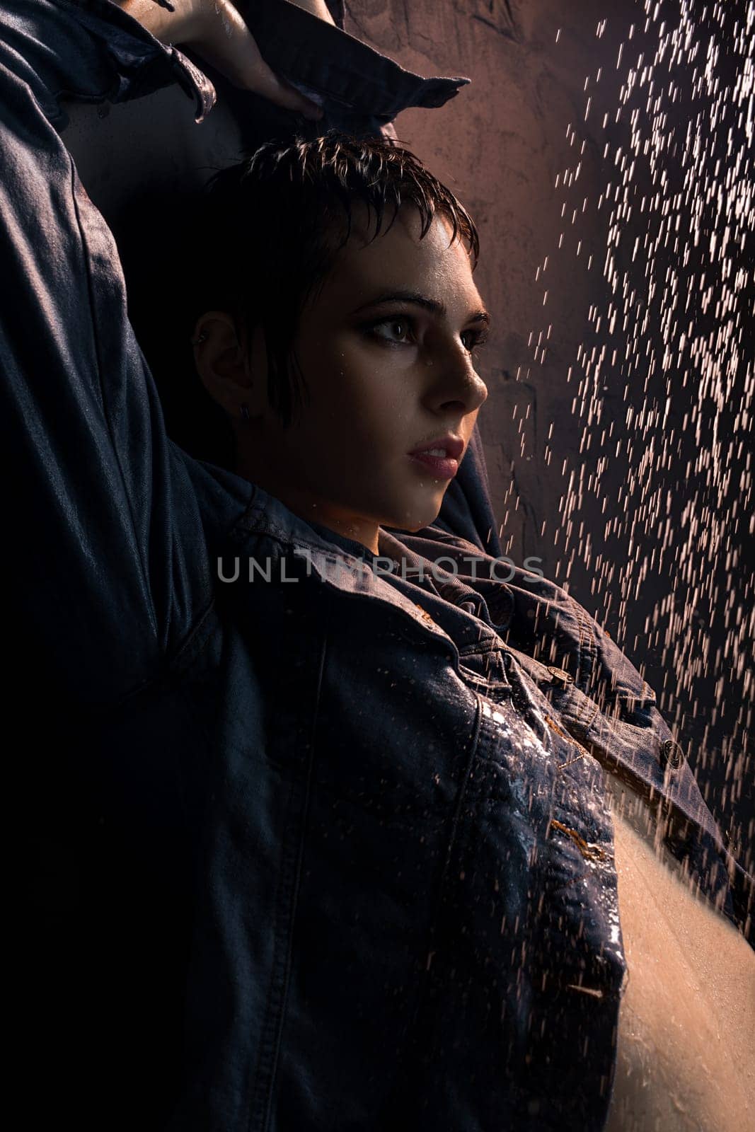 Brunette in wet jeans shirt standing under water in dark room profile portrait
