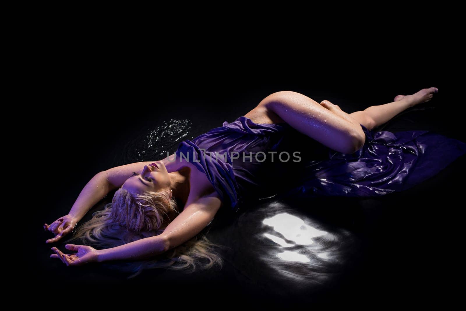 Sexy blonde wrapped in purple cloth lying in water in the dark room high angle view