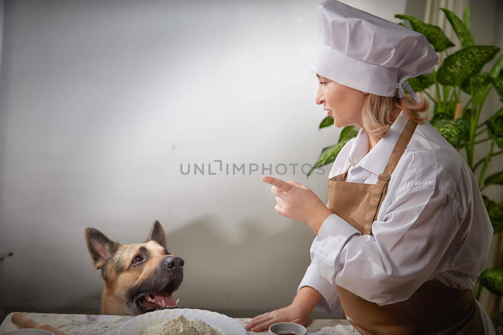 An elderly woman dressed as a cook shares a light-hearted moment with her shepherd dog while preparing flour-based dishes.