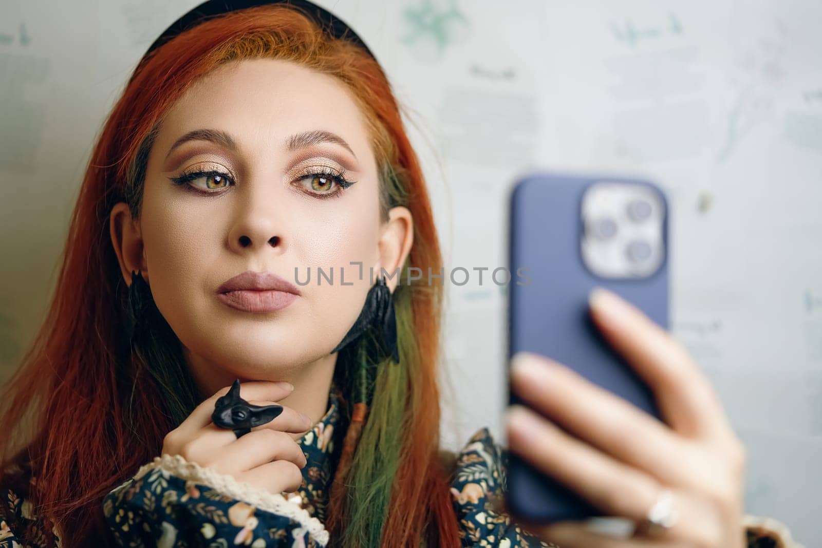 Beautiful girl takes a selfie on the front camera of a modern smartphone. Female blogger films herself for a fashion and makeup blog. Red-haired woman with jewelry in Gothic style. Witch aesthetic.