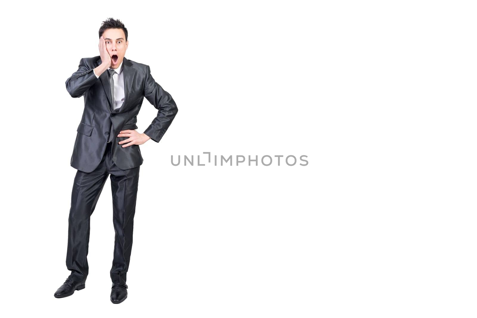 Full body of astonished young male manager in formal clothes touching cheek and looking at camera with opened mouth while standing against white background with hand on waist