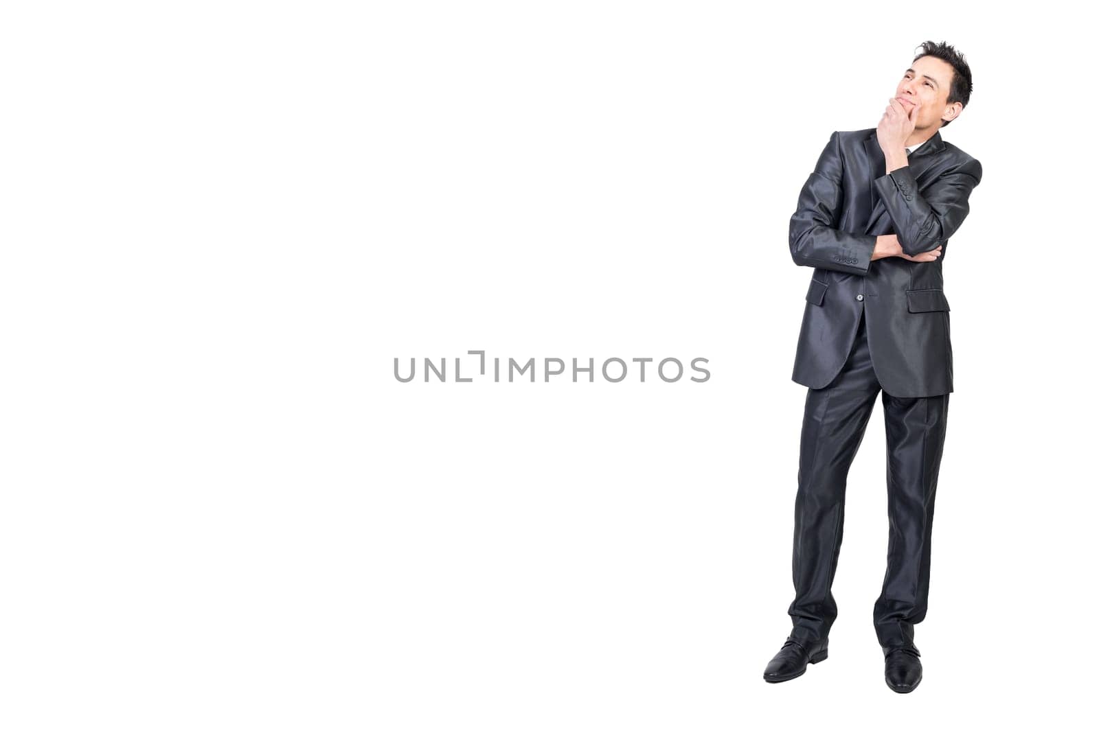 Full body of elegant young male manager with dark hair in classy suit touching chin and looking away dreamily in white studio