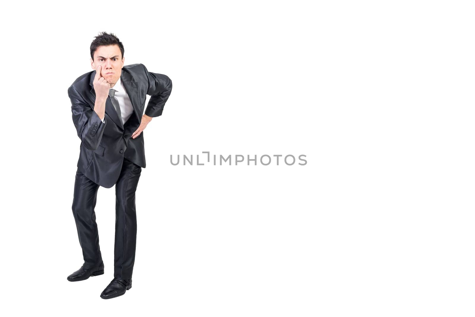 Full body of serious young male manager with dark hair in formal suit pointing at eye and gazing at camera while standing against white background with hand on waist