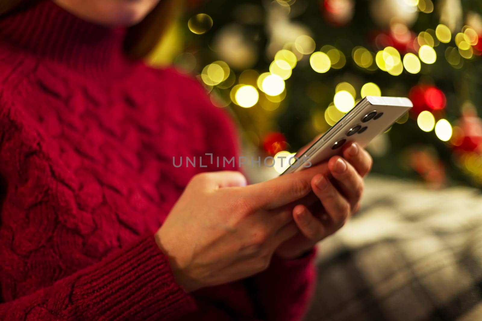 Woman holding a smartphone while sitting near a Christmas tree decorated with lights, wearing a red sweater. Holiday atmosphere depicting digital communication and celebration.