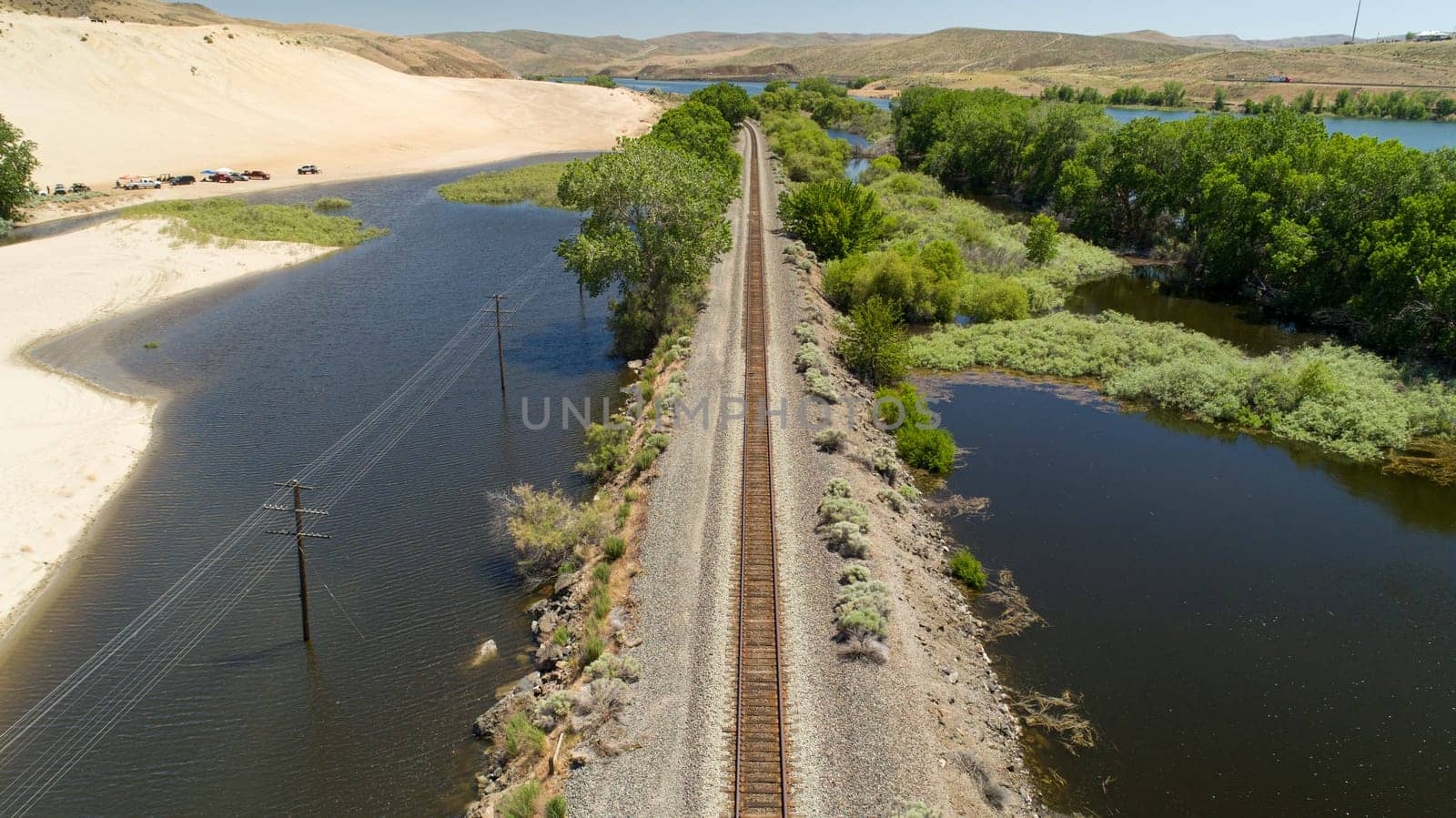 Aerial shot showcasing a railway track running through a lush, green landscape, bordered by serene water bodies and sand dunes extending into the horizon.