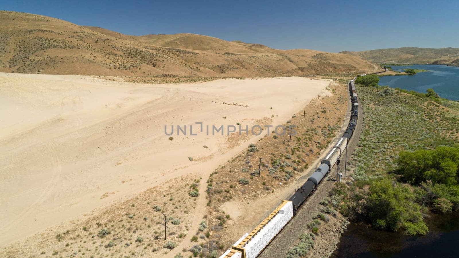 Aerial view of a freight train winding through a vast desert landscape beside a tranquil river. The scene captures the essence of solitude and adventure in a remote, natural environment.