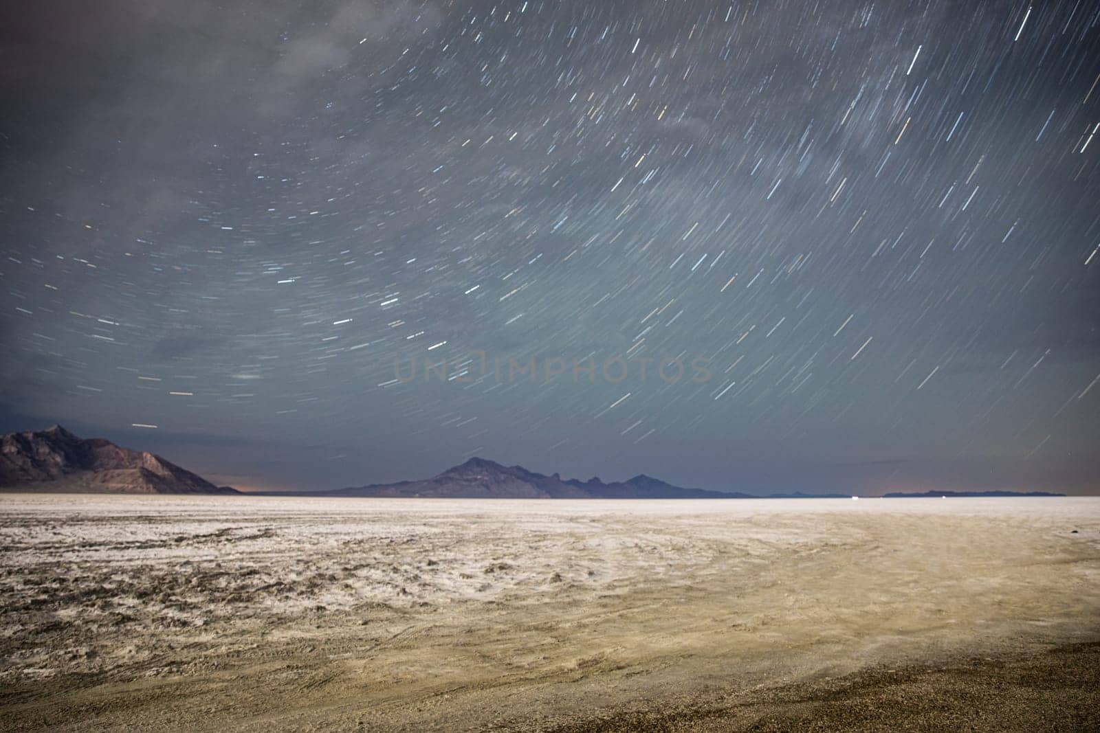 A mesmerizing view of the starry night sky with long exposure over a wide, barren desert. The serene desert terrain beautifully contrasts the dynamic star trails in the sky.