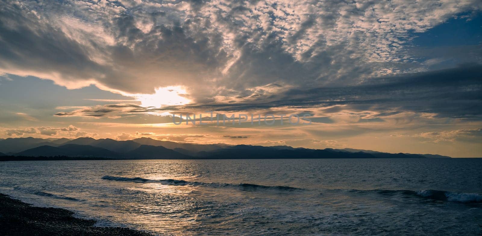 Waves gently lap the shore as the sun sets behind mountains, casting warm hues across the expansive sky.