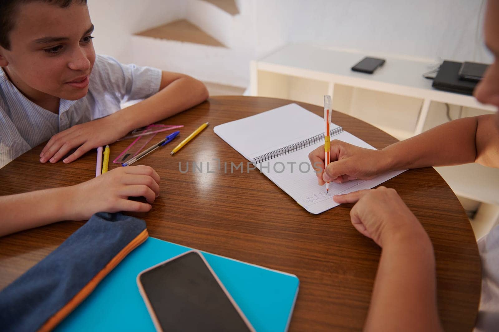 Two children are sitting at a table engaged in a study session. One is writing in a notebook while the other watches attentively. Pencils and school supplies are scattered on the table.