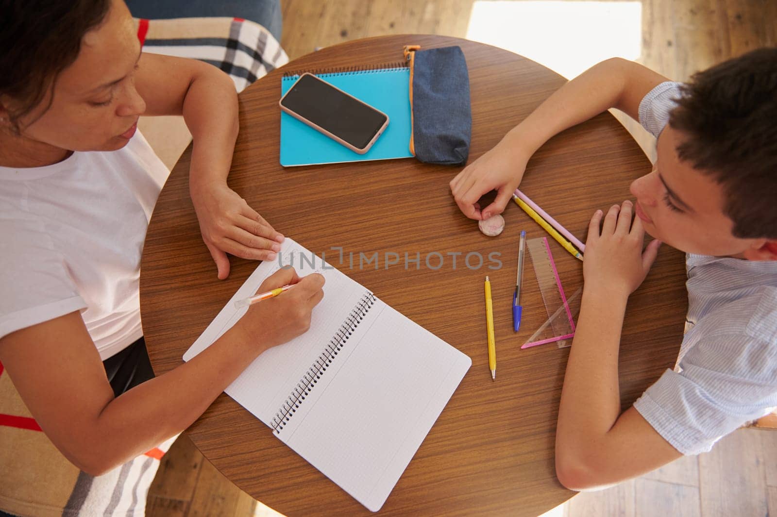 A mother assists her son with homework at a round wooden table. Pencils, a notebook, and a smartphone are on the table, fostering a supportive and educational environment.