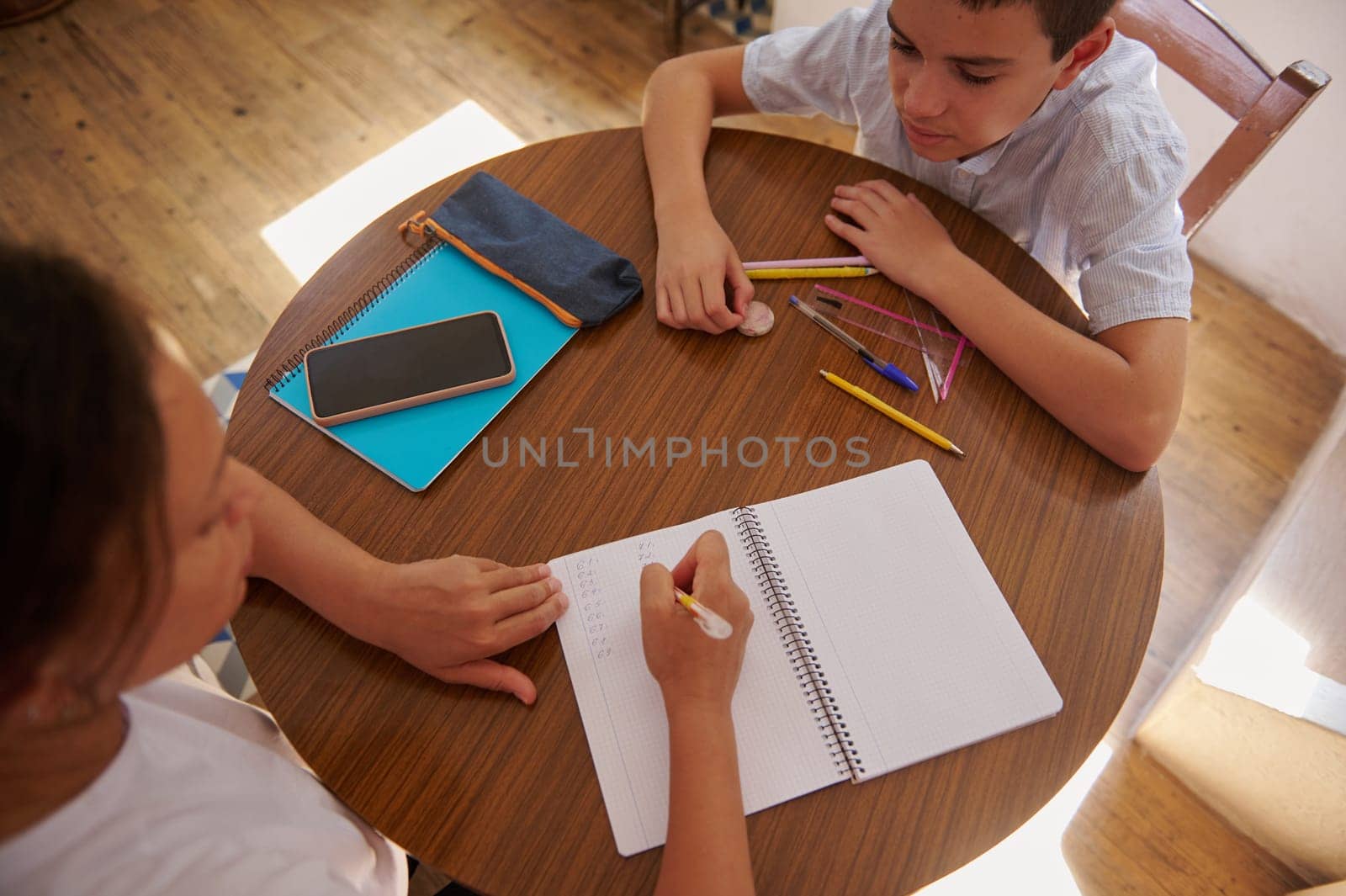 A mother helps her son with homework, writing in a notebook on a wooden table. They are surrounded by school supplies and focusing on the task, creating a supportive learning environment.