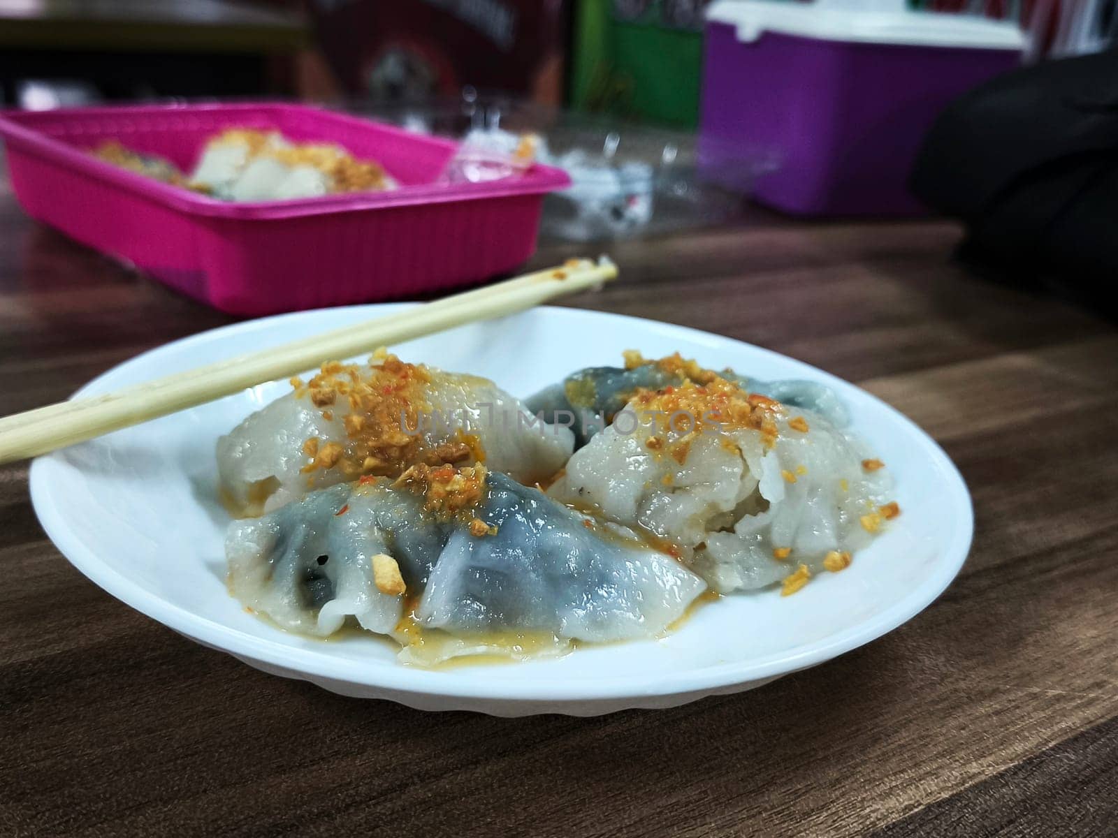 A plate of Choipan cakes from Indonesia sits on a wooden table. The cakes are white with green accents and are topped with a red sauce. Chopsticks are placed on the plate.