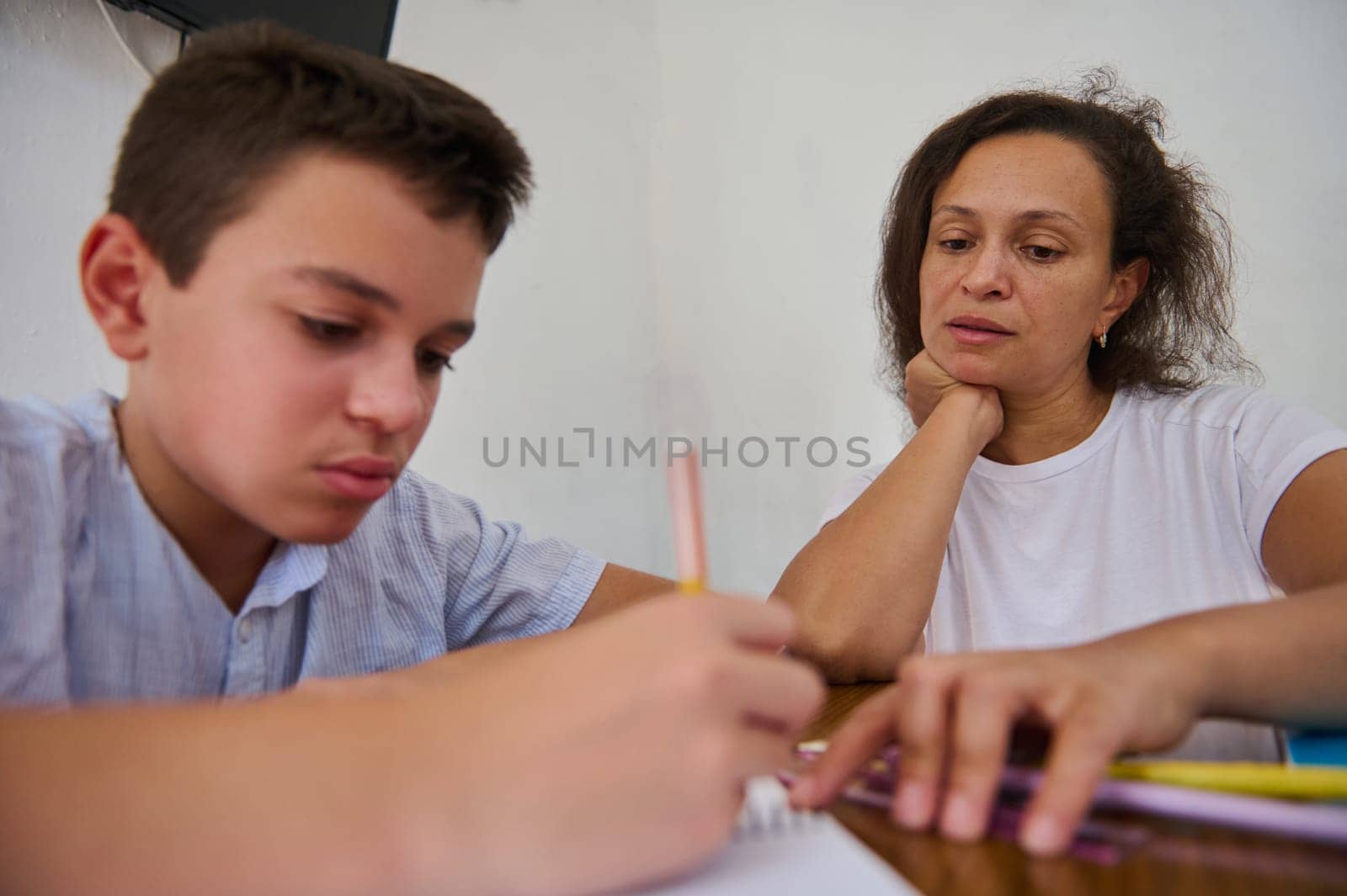 A mother attentively assists her son with his homework at the kitchen table. The boy focuses on writing while the mother guides him, displaying a scene of learning and support at home.