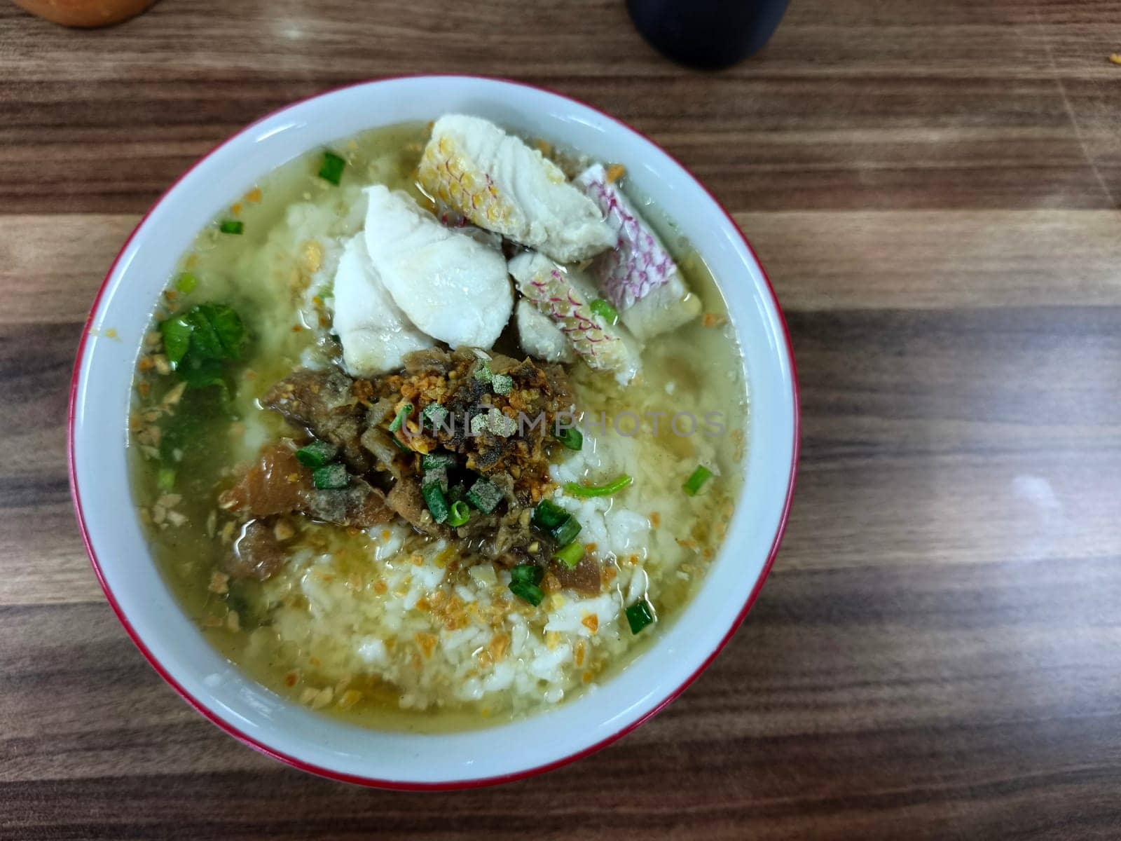 A bowl of Bubur Ikan Singkawang sits on a wooden table. The porridge is topped with chunks of fish and a red sauce. Chopsticks and other dishes are visible in the background.
