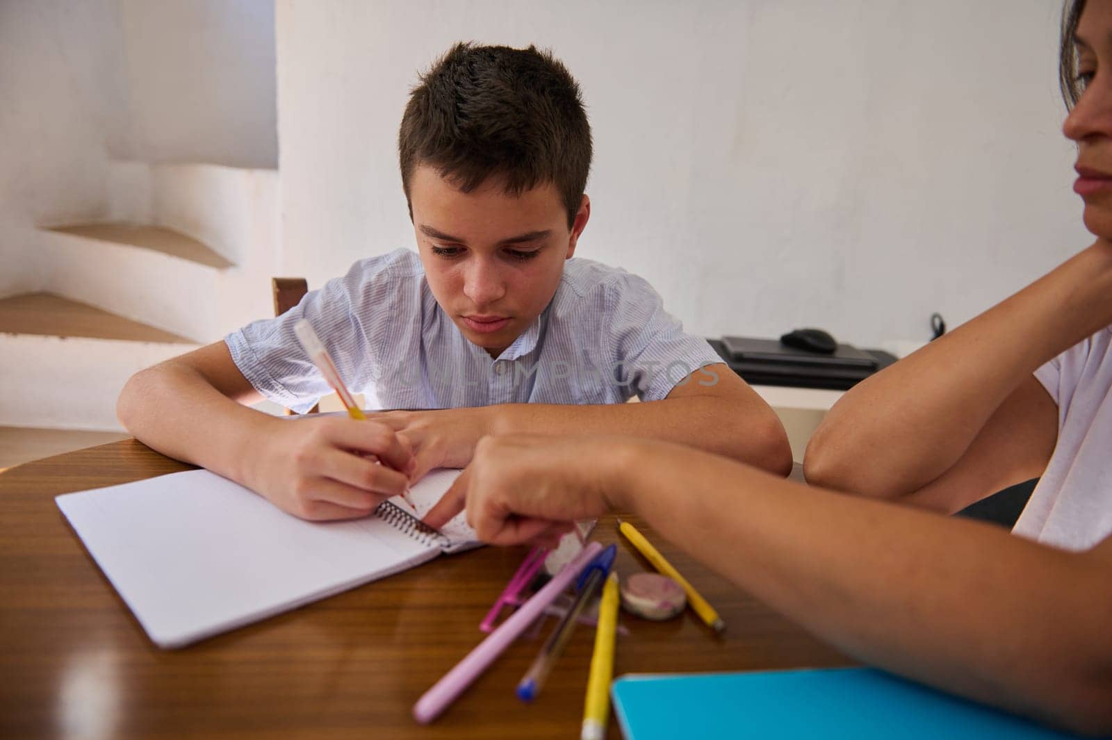 A young boy focuses intensely on his homework as an adult guides him, surrounded by colorful stationery on a wooden table.