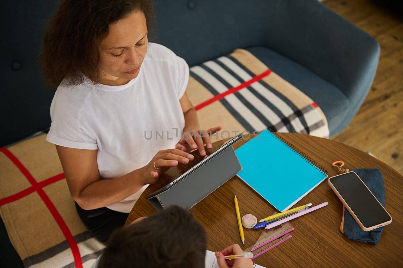 A woman assists her child with homework using a digital tablet at a cozy home setting, fostering learning.