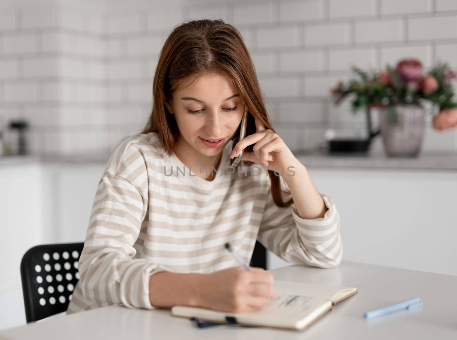Young Girl Talks On Phone And Makes Notes In Notebook At Home
