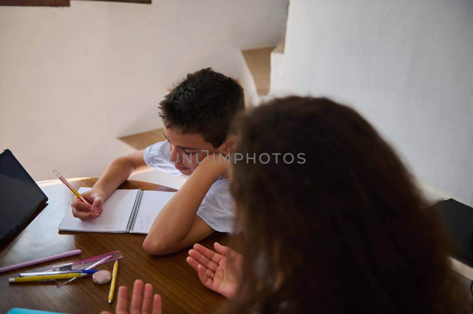 A young boy sits at a wooden table, focused on writing in a notebook, as a woman assists him with his homework. Various school supplies are scattered on the table.
