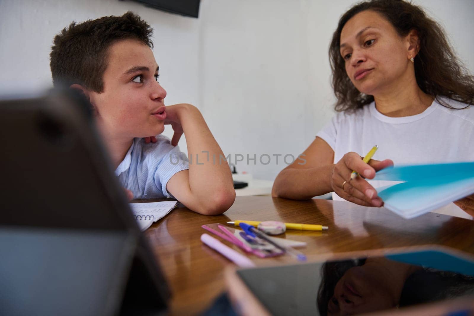 A mother helps her son with homework, offering guidance and support. The interaction promotes learning, education, and family bonding. Pens and notebooks are scattered on the table.